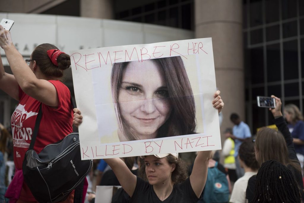 Image of a woman in a black shirt holding aloft a large photo portrait of Heather Heyer with the words "Remember Her" above and "Killed By Nazi" below the photo. To her left is another woman in a red "Dump Trump" shirt.