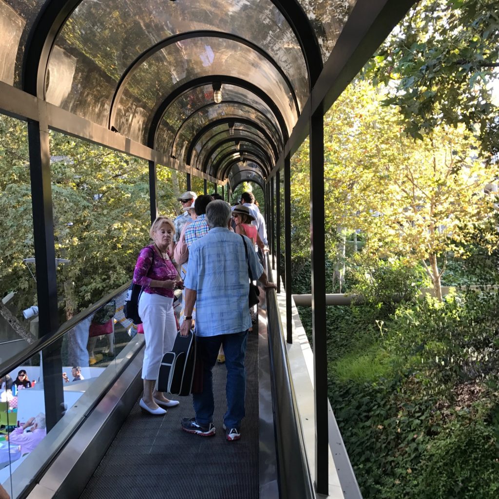 Image is of the Hollywood Bowl escalator, which is flat and operates as a moving walkway. It has a clear canopy over top and several concertgoers are standing in conversation as they ride.
