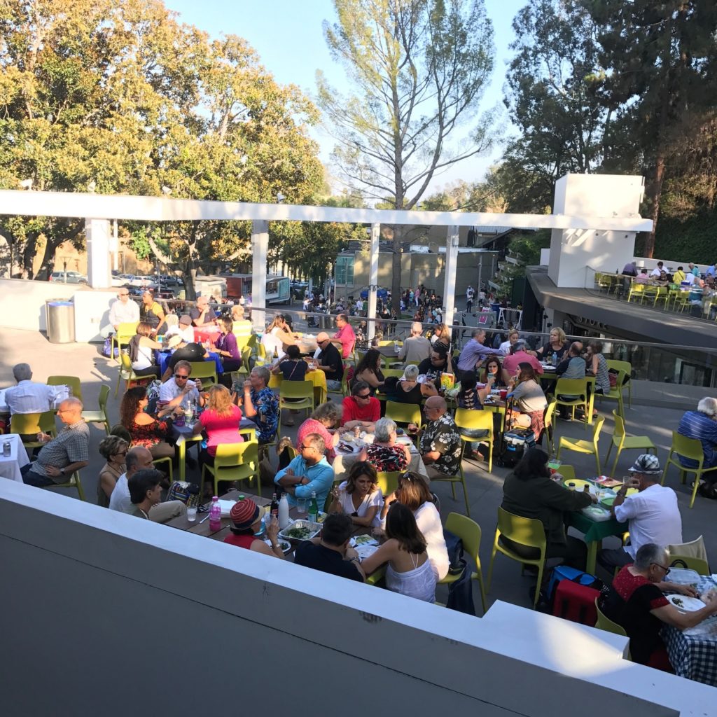 Image of the picnic area at the Hollywood Bowl with concertgoers seated in chartreuse-colored chairs around small tables in dappled golden sunlight.