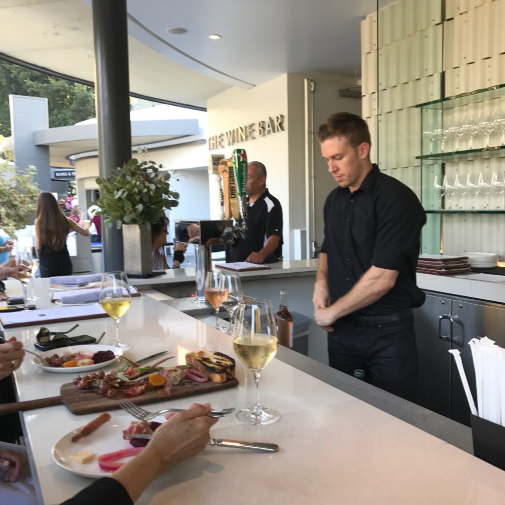 Image of a bartender behind the counter at the Hollywood Bowl Wine Bar. On the bar is a wood board featuring a selection of charcuterie, two glasses of chilled white wine, and the filled plates of two diners.