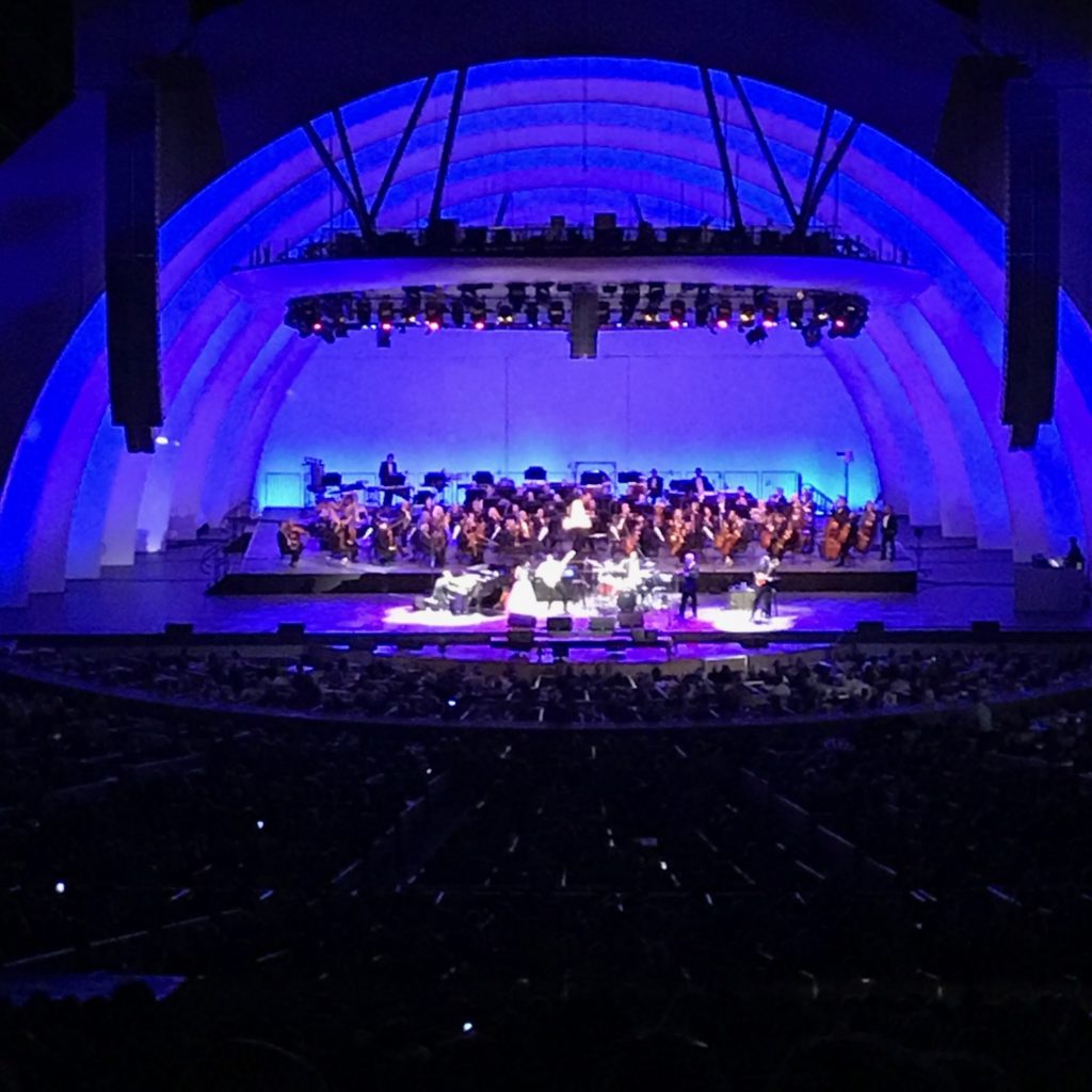 Image is of several performers downstage in the center of the Hollywood Bowl shell with the orchestra seated upstage. The shell is lit with deep blue light and white spotlights on the performers.